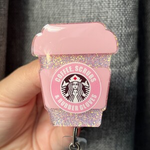 May include: Pink glitter coffee cup with a Starbucks logo and the text "Coffee, Scrubs & Rubber Gloves". The cup is attached to a pink glitter retractable badge holder.