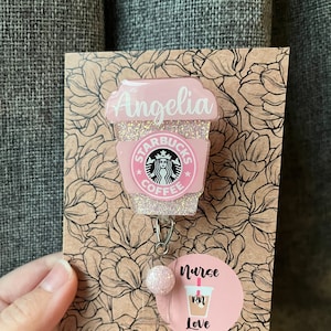 May include: Pink glitter coffee cup with the Starbucks logo and the name "Angelia" on the side. The cup is attached to a pink glitter badge reel with a pink glitter bead. A round sticker with the text "Nurse RN Love" is attached to the reel.