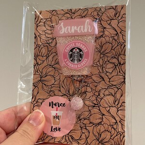 May include: Pink and white coffee cup charm with glitter and the text "Sarah, Coffee, Scrubs & Rubber Gloves". The charm is attached to a pink and white circle with the text "Nurse RN Love".