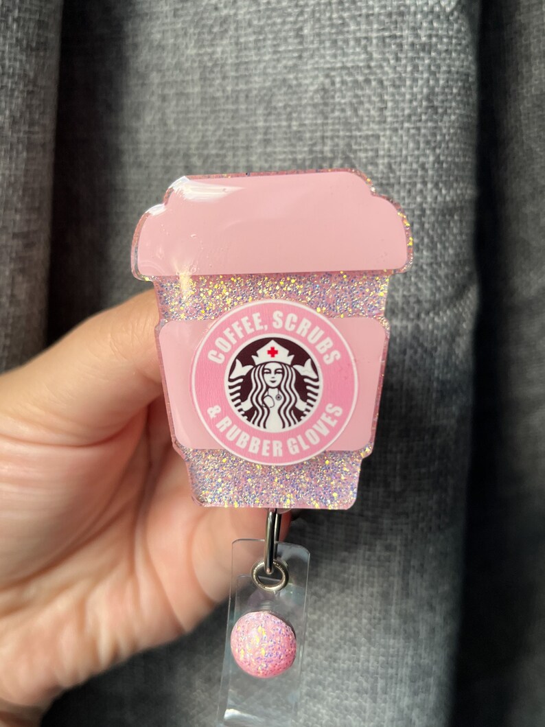 May include: Pink glitter coffee cup with a Starbucks logo and the text "Coffee, Scrubs & Rubber Gloves". The cup is attached to a pink glitter retractable badge holder.