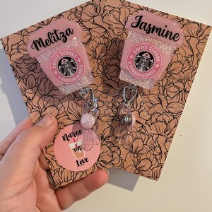 May include: Two pink and white glitter coffee cup themed retractable badge holders with the text "Coffee, Scrubs & Rubber Gloves" and the names "Melitza" and "Jasmine". The badge holders are on a floral patterned background with a pink circle sticker that says "Nurse RN Love".