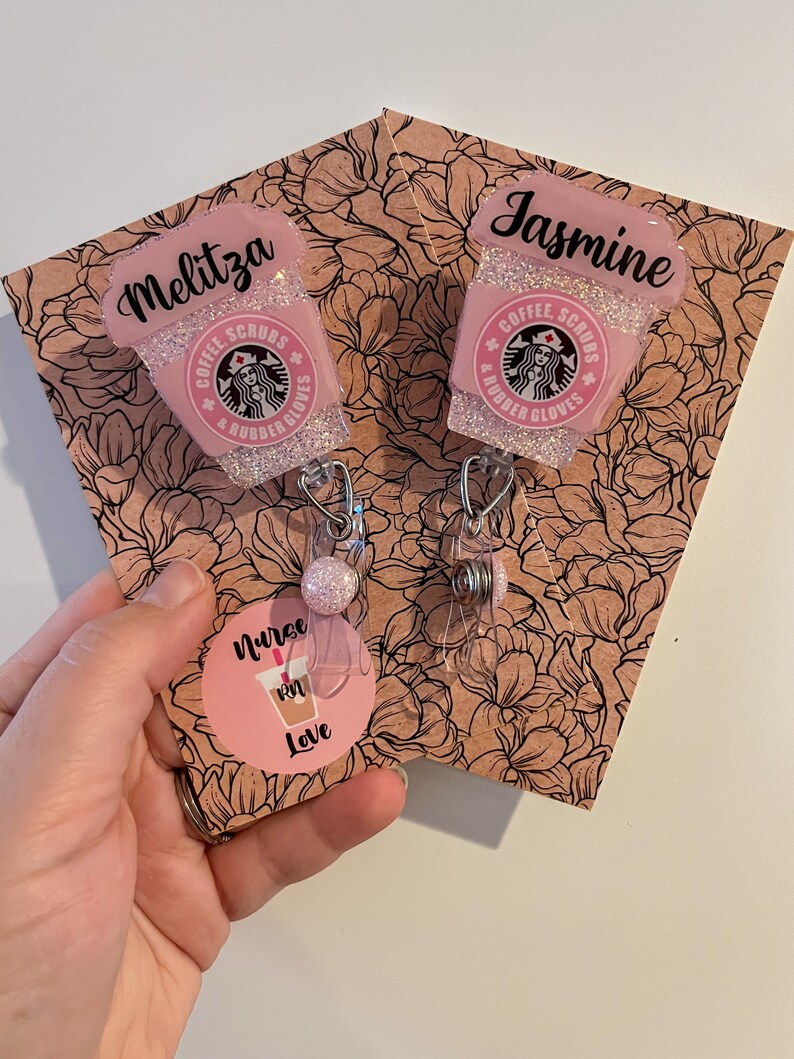 May include: Two pink and white glitter coffee cup themed retractable badge holders with the text "Coffee, Scrubs & Rubber Gloves" and the names "Melitza" and "Jasmine". The badge holders are on a floral patterned background with a pink circle sticker that says "Nurse RN Love".