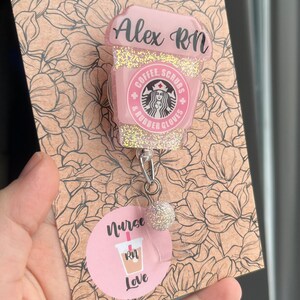 May include: Pink and gold glitter coffee cup charm with the text "Alex RN", "Coffee, Scrubs & Rubber Gloves" and a Starbucks logo. The charm is attached to a retractable badge holder with a pink and gold glitter ball. The charm is displayed on a brown card with a floral pattern and the text "Nurse RN Love".