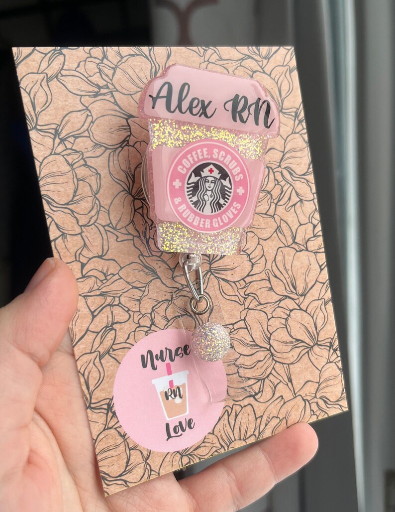 May include: Pink and gold glitter coffee cup charm with the text "Alex RN", "Coffee, Scrubs & Rubber Gloves" and a Starbucks logo. The charm is attached to a retractable badge holder with a pink and gold glitter ball. The charm is displayed on a brown card with a floral pattern and the text "Nurse RN Love".