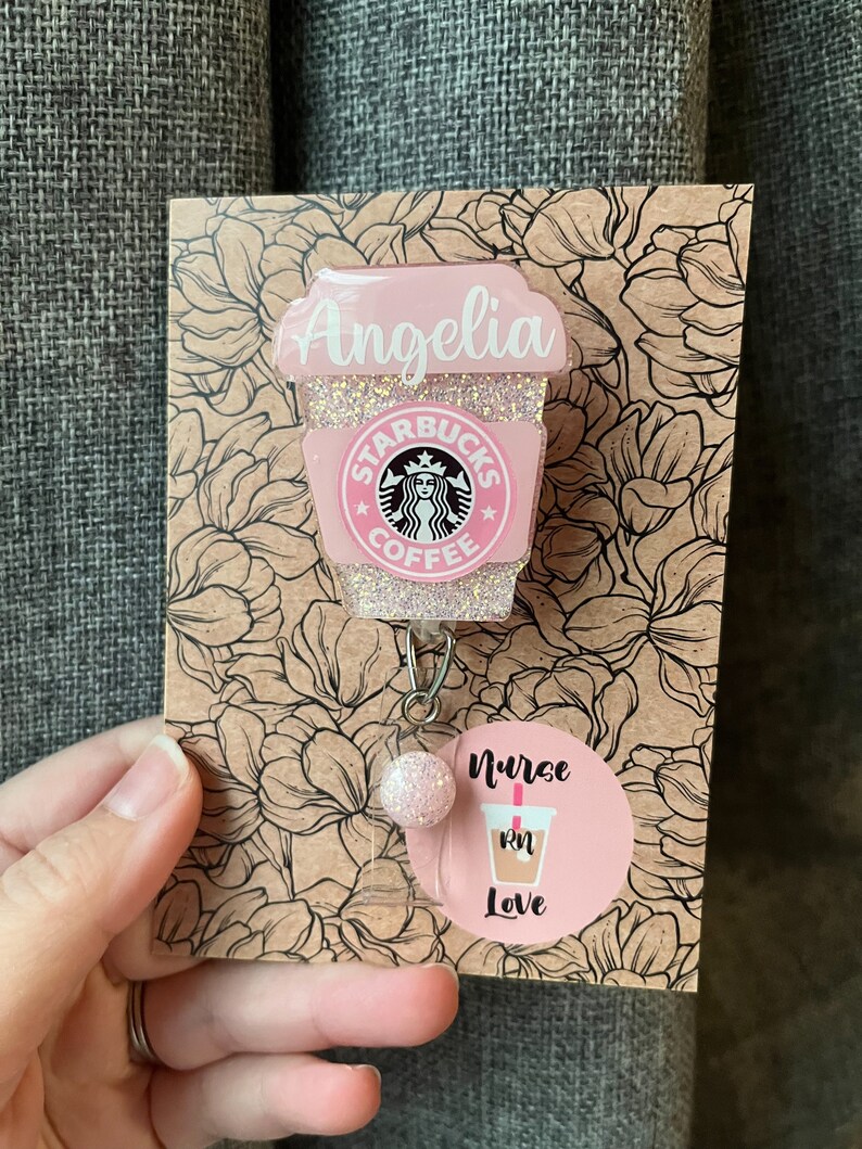 May include: Pink glitter coffee cup with the Starbucks logo and the name "Angelia" on the side. The cup is attached to a pink glitter badge reel with a pink glitter bead. A round sticker with the text "Nurse RN Love" is attached to the reel.