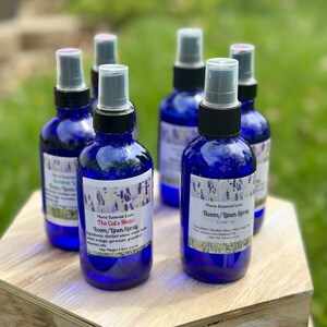 May include: Four blue glass bottles with spray tops and labels. The labels are white with black text and lavender flowers. The text on the labels reads "Moms Essential Love", "The Cat's Meow", "Room/Linen Spray", and "Cover Up".
