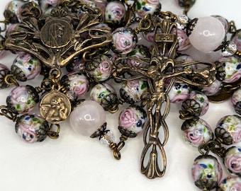 Peut inclure: Un chapelet composé d'éléments en métal couleur bronze antique, incluant un crucifix et une médaille centrale. La chaîne est formée de perles en verre blanc peintes de roses roses et de feuilles vertes, alternées avec des perles rondes rose pâle.