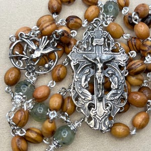 Pode incluir: Um rosário de prata com contas de madeira de oliveira e um crucifixo de prata com uma figura de Jesus Cristo. O crucifixo é adornado com detalhes intrincados e dois anjos. O rosário também inclui um pingente de prata com uma pomba.