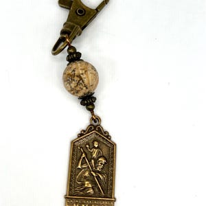 May include: A bronze-colored keychain featuring a religious pendant. The pendant depicts a saint carrying a child, with a decorative border. The keychain includes a patterned bead and a clasp.