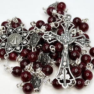 Op de afbeelding: Een rozenkrans met dieprode kralen en sierlijk zilverkleurig metaalwerk. Het middelpunt is een gedetailleerd kruisbeeld en de ketting bevat een decoratieve hanger met een religieuze figuur. De kralen zijn rond en gelijkmatig verdeeld.