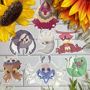 Puede incluir: Un conjunto de ocho pegatinas con personajes del videojuego Hollow Knight. Las pegatinas están impresas en papel blanco y presentan una variedad de colores, incluyendo rojo, azul, verde y negro. Las pegatinas son de diferentes tamaños y formas, y todas están diseñadas para parecerse a los personajes del juego.