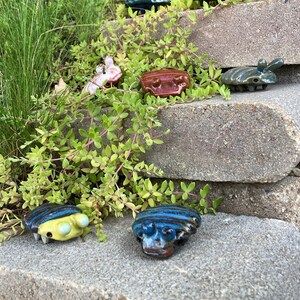 May include: A collection of ceramic figurines in various colors and designs, resembling small creatures with multiple legs. The figurines are arranged on a set of stone steps, with green foliage surrounding them.