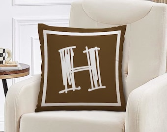 May include: A brown armchair with a white cushion featuring a white letter H on a brown background. The cushion has a white border.