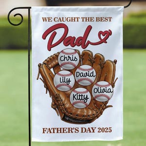 Puede incluir: Una bandera de jardín blanca con un guante de béisbol y pelotas de béisbol. El texto dice "WE CAUGHT THE BEST Dad" en escritura roja. Las pelotas de béisbol tienen nombres: Chris, Lily, David, Kitty y Olivia. El texto inferior dice "FATHER'S DAY 2025".
