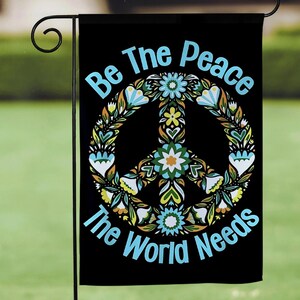Könnte beinhalten: Eine schwarze Gartenflagge mit einem Peace-Zeichen, das von Blumen umgeben ist, und dem Text "Be The Peace The World Needs" in Blau.
