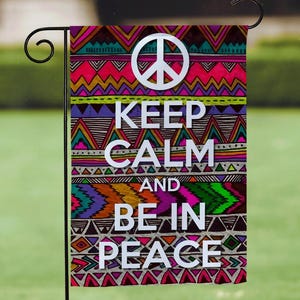 May include: A colorful garden flag with a peace symbol and the words "KEEP CALM AND BE IN PEACE" in white. The flag features a vibrant geometric pattern in shades of pink, purple, orange, and blue. The flag is displayed outdoors in a garden setting.