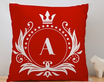 May include: Red cushion with a white decorative design, including a crown and the letter "A". The square cushion is placed on a wooden surface. The design is symmetrical.