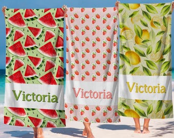 May include: Three beach towels with fruit patterns are held up on a sandy beach. One towel has watermelon slices on a green background, another has strawberries on pink, and the last has lemons on a yellow background. Each towel has the name "Victoria" on it.