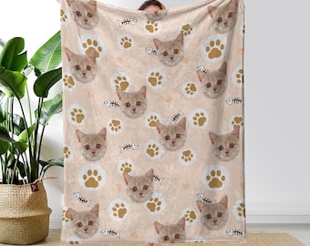 May include: A soft, plush blanket featuring a repeating pattern of cat faces and paw prints on a light beige background. The cat faces are all different, but they are all smiling and have a playful expression.