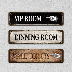 Puede incluir: Tres letreros de metal de estilo vintage con bordes oxidados y texto blanco. El letrero superior es negro con texto blanco que dice "VIP ROOM" y una mano señalando. El letrero del medio es blanco con texto negro que dice "DINNING ROOM". El letrero inferior es marrón con texto blanco que dice "MALE TOILETS" y una mano señalando.