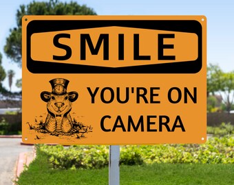May include: Orange and black sign with the text "Smile You're On Camera" and a cartoon image of a groundhog wearing a top hat and holding a pocket watch.