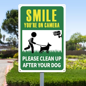 Könnte beinhalten: Ein rechteckiges grünes und weißes Schild mit dem gelben Text "SMILE YOU'RE ON CAMERA". Darunter eine Grafik einer Person, die einen schwarzen Hund ausführt, und der Text "PLEASE CLEAN UP AFTER YOUR DOG".