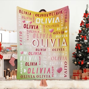 Op de afbeelding: Een gepersonaliseerde deken met de naam "OLIVIA" in witte en roze letters. De deken heeft een gradiënt achtergrond van geel, roze en groen. Harten zijn verspreid over het ontwerp. Een kerstboom staat op de achtergrond.