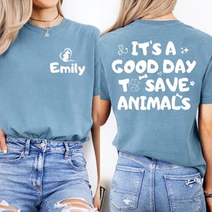 May include: A dusty blue short-sleeve t-shirt featuring a white graphic on the front left chest that reads Emily with a heart and paw print icon. The back displays the white text It's a good day to save animals with stethoscope and paw icons.