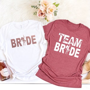 May include: Two t-shirts, one white and one coral pink, with the text "BRIDE" and "TEAM BRIDE" in white and rose gold lettering. The shirts feature a silhouette of a woman dancing on a pole.