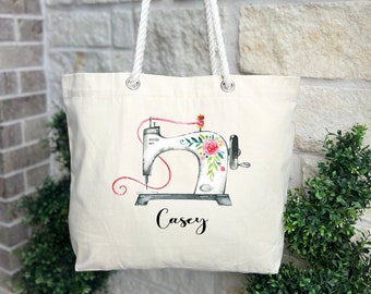 May include: A white canvas tote bag with a floral design of a vintage sewing machine and the name "Casey" printed on the front. The bag has white rope handles.
