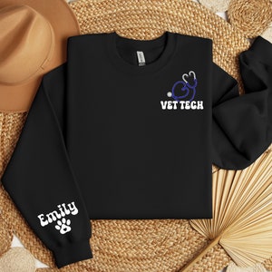May include: Black sweatshirt with a white paw print and the name "Emily" on the left sleeve. The front of the sweatshirt has a blue stethoscope with a heart design and the text "VET TECH" in white.
