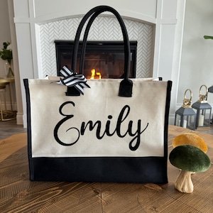 May include: A canvas tote bag with black handles and a black bottom panel. The name "Emily" is printed in a cursive font on the front. A black and white striped bow is attached to one of the handles.