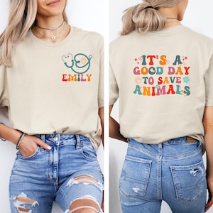 May include: Two women wearing beige t-shirts with different designs. The shirt on the left has a stethoscope and the name "Emily" printed on it. The shirt on the right has the text "It's a good day to save animals" printed on it in colorful letters with paw prints and bone icons. Both women are wearing blue jeans.
