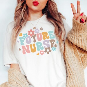 May include: A white t-shirt with the text "FUTURE NURSE" in colorful, retro-style lettering and flowers. The shirt is being worn by a person with long blonde hair.
