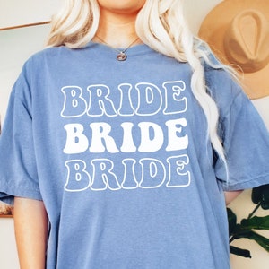 Pode incluir: Uma camiseta oversized azul claro com a palavra "BRIDE" repetida tr&ecirc;s vezes em uma fonte retr&ocirc; em branco.