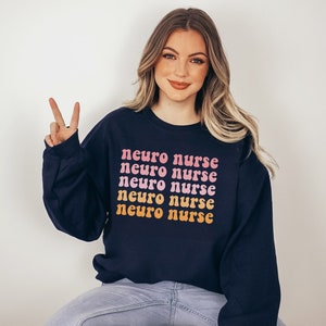 May include: A navy blue sweatshirt with the text "neuro nurse" repeated five times in a rainbow gradient. The text is in a retro font style.