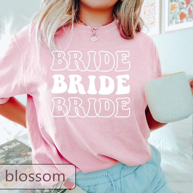 Pode incluir: Uma camiseta rosa com a palavra "BRIDE" escrita tr&ecirc;s vezes em um estilo de fonte retr&ocirc;.