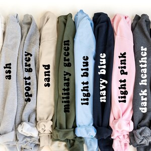 May include: A row of ten sweatshirts in different colors, including white, ash, sport grey, sand, military green, light blue, navy blue, light pink, dark heather, and black. Each sweatshirt has the color name printed on the fabric.