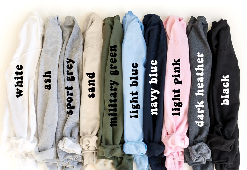 May include: A row of ten sweatshirts in different colors, including white, ash, sport grey, sand, military green, light blue, navy blue, light pink, dark heather, and black. Each sweatshirt has the color name printed on the fabric.
