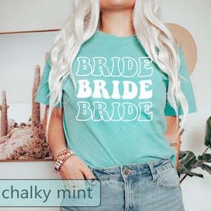 Pode incluir: Uma camiseta azul turquesa com a palavra "BRIDE" repetida tr&ecirc;s vezes em uma fonte retr&ocirc; em branco.