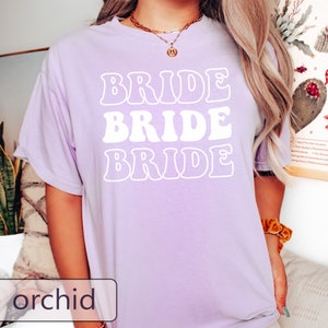 Pode incluir: Uma camiseta de cor lavanda com a palavra "BRIDE" repetida tr&ecirc;s vezes em um estilo de fonte retr&ocirc;. O texto &eacute; branco.