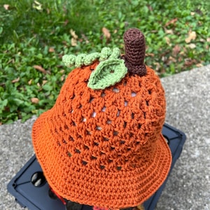 May include: An orange crochet bucket hat shaped like a pumpkin, complete with a brown stem and green leaves. The hat is made of a textured yarn and has a wide brim. Perfect for fall or Halloween.
