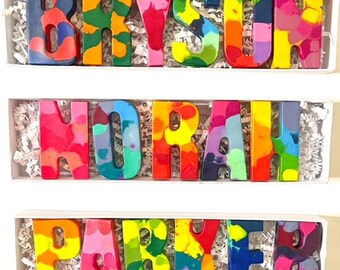 May include: Three sets of colourful crayon letters spelling out the names Bryson, Norah, and Parker. The letters are arranged in a row in a white box with white shredded paper.