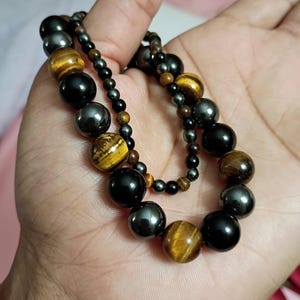 Puede incluir: Dos pulseras de cuentas con una mezcla de cuentas esféricas pulidas. Una pulsera consta de cuentas más grandes en negro, gris metálico y ojo de tigre marrón dorado. La segunda pulsera presenta cuentas más pequeñas en negro, gris metálico y ojo de tigre.