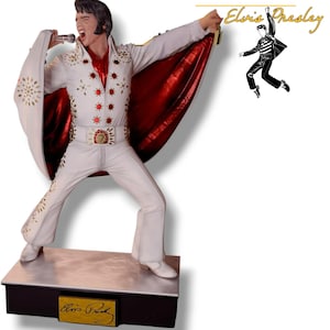 May include: A white statue of Elvis Presley in a performance pose, wearing a white jumpsuit with gold embellishments and a red-lined cape. The statue holds a microphone and is on a black base with a gold nameplate that reads "Elvis Presley".