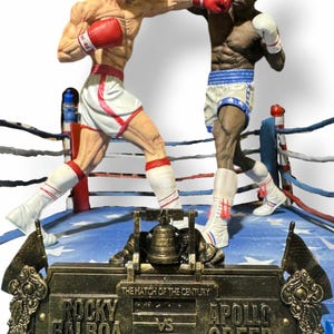 Puede incluir: Un diorama de ring de boxeo con figuras de Rocky Balboa y Apollo Creed. El ring está decorado con un patrón de bandera estadounidense azul y roja. Una placa dorada dice "The Match of the Century" con "Rocky Balboa" y "Apollo Creed" a cada lado.
