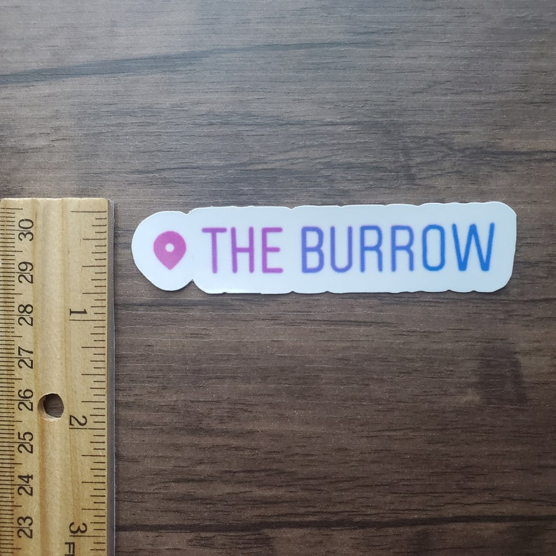K&ouml;nnte beinhalten: Ein wei&szlig;er Aufkleber mit einem rosa Standort-Pin und dem Text "THE BURROW" in Lila und Blau.