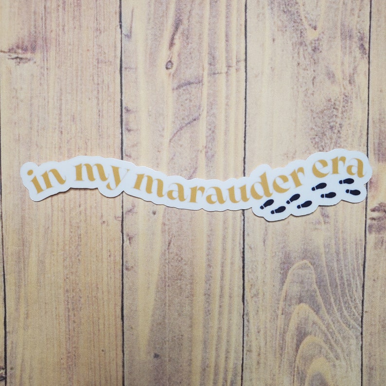 May include: A white sticker with the text "in my marauder era" in gold lettering and a trail of black footprints.