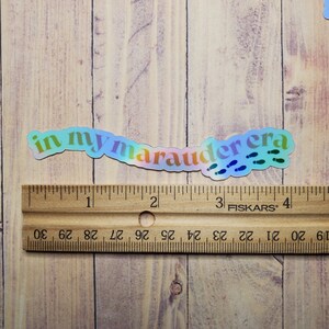 May include: A holographic sticker with the text "in my marauder era" in rainbow colors with blue footprints.