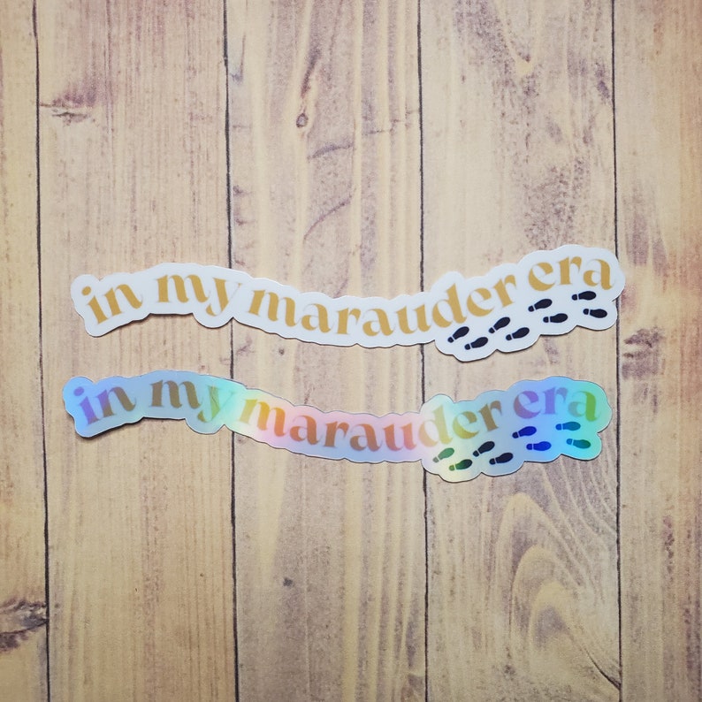 May include: Two iridescent stickers with the text "in my marauder era" and a series of footprints. The stickers are on a woodgrain background.